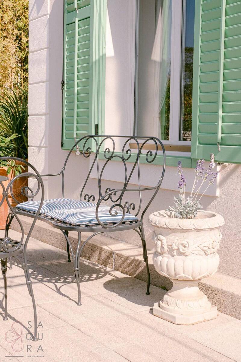 Detailansicht einer schmiedeeisernen Bank auf der Terrasse mit Blumentopf und Fensterläden.