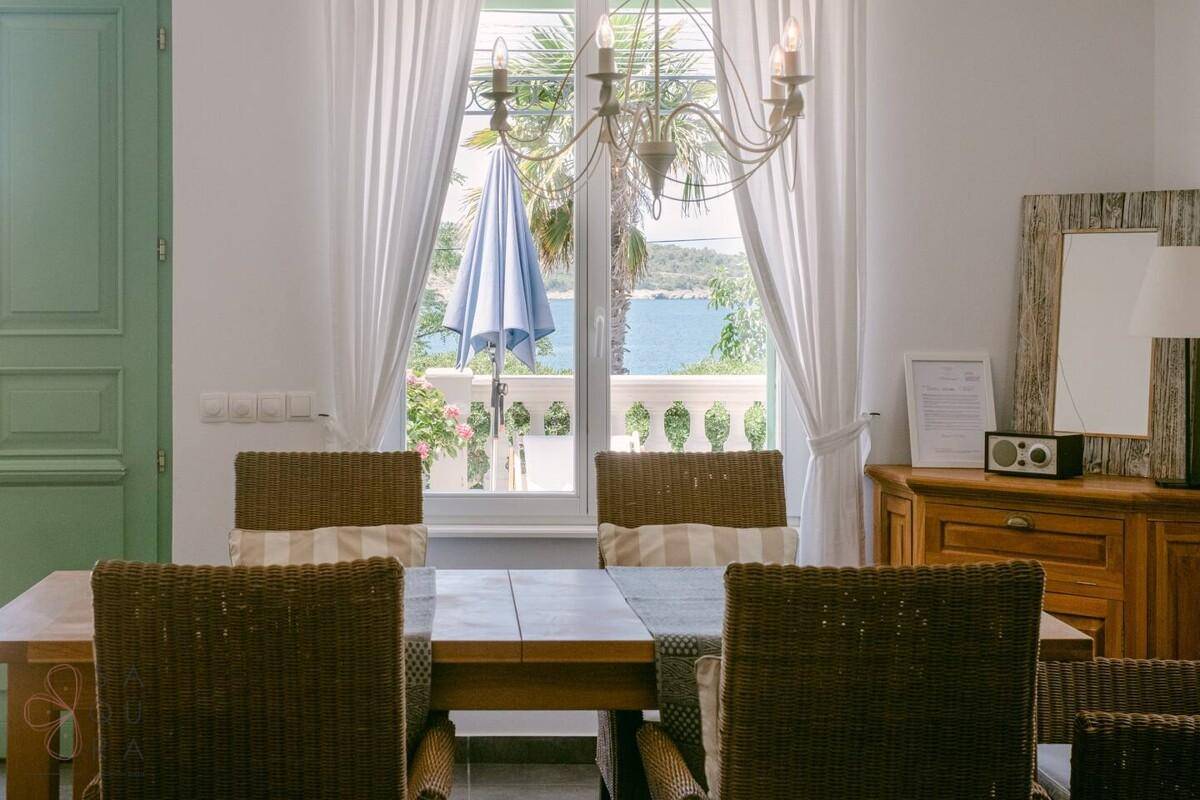 Heller Essbereich mit Holztisch, Korbstühlen und Meerblick durch das Fenster.