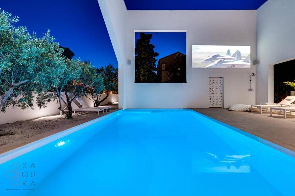 Beleuchteter Poolbereich bei Nacht mit Projektion an der Wand und mediterranen Olivenbäumen.