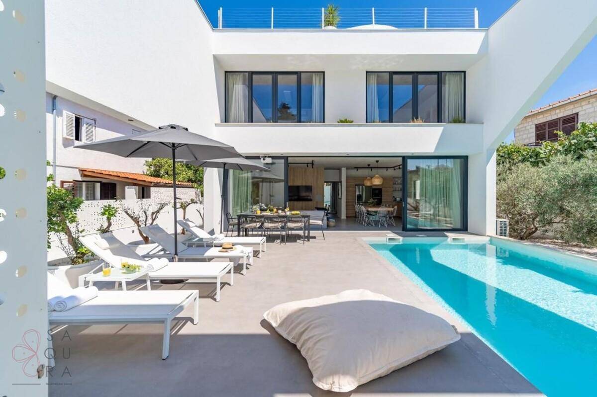 Moderne Villa mit großem Pool, Sonnenterrasse und Essbereich im Freien unter Sonnenschirmen.