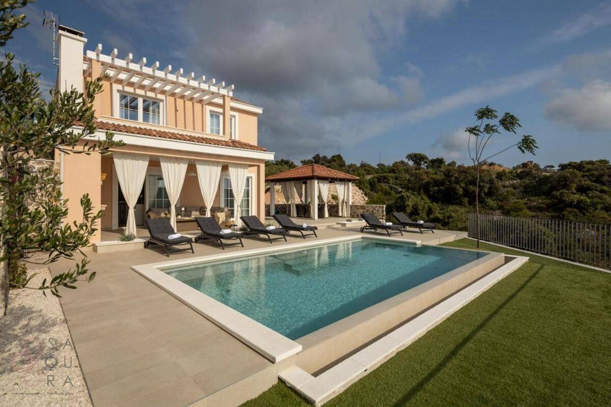 Luxuriöse Villa mit großem Poolbereich und Liegestühlen unter blauem Himmel, umgeben von mediterraner Landschaft.