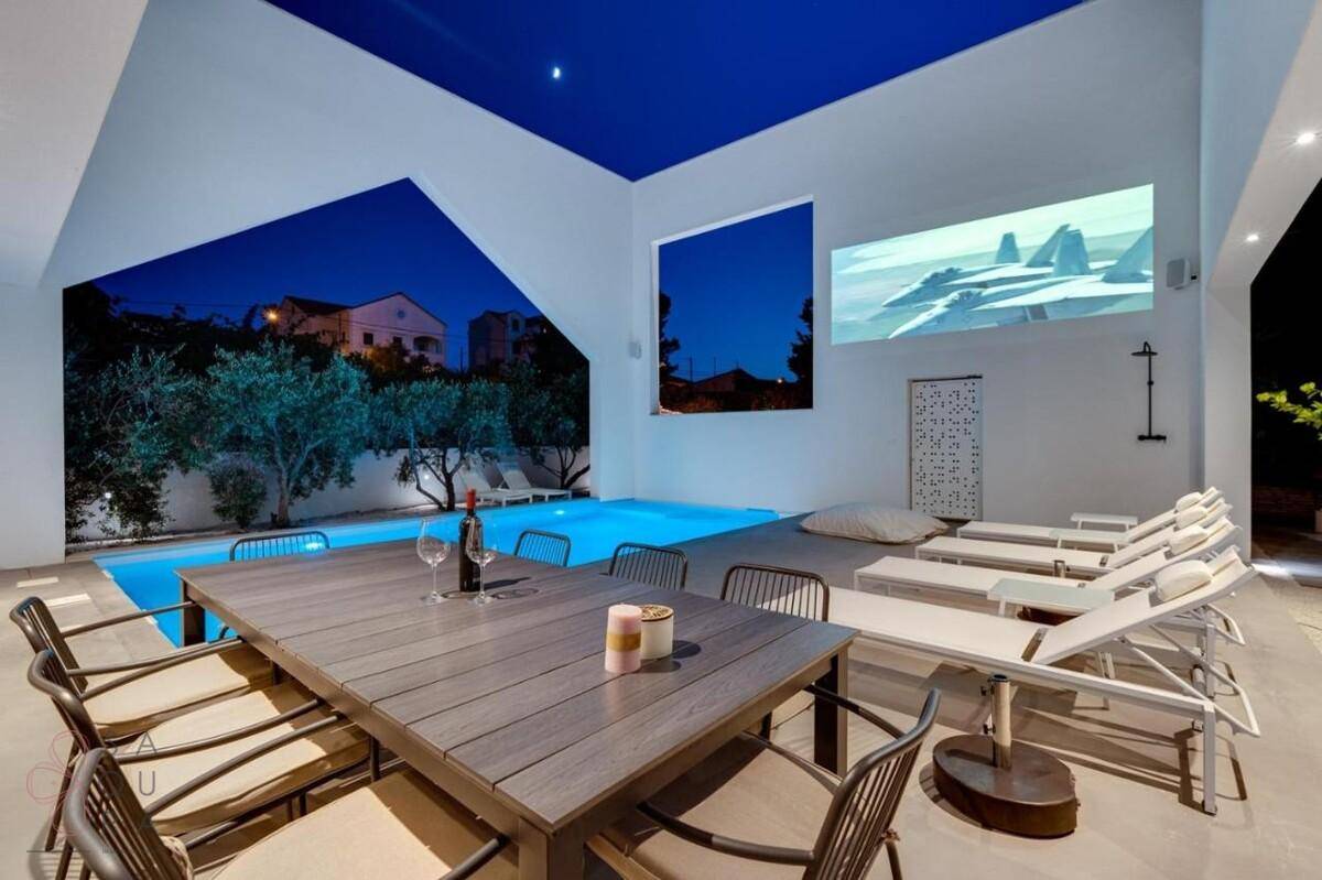 Beleuchteter Poolbereich mit Essbereich im Freien und Projektion an der Wand bei Nacht.