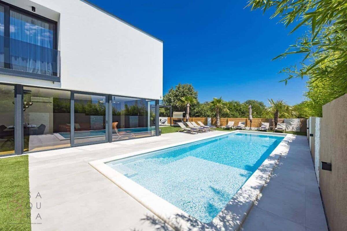 Sonniger Poolbereich mit klarem Wasser, umgeben von Grün und moderner Architektur.