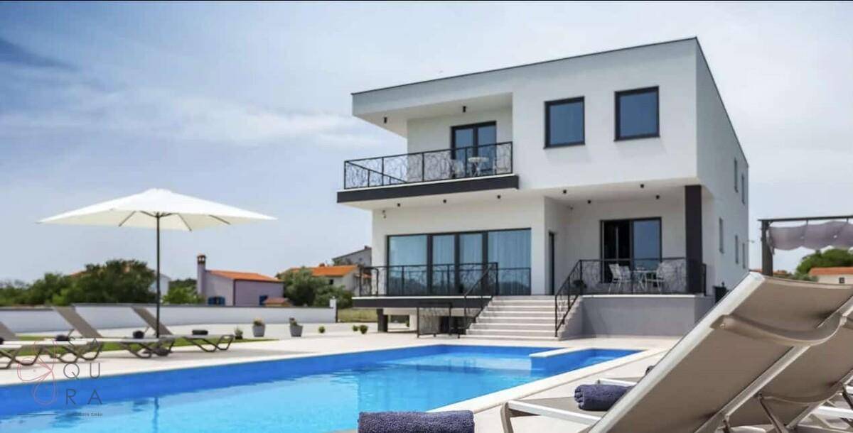 Ansicht der modernen Villa mit großem Poolbereich und zahlreichen Liegestühlen unter hellem Himmel.