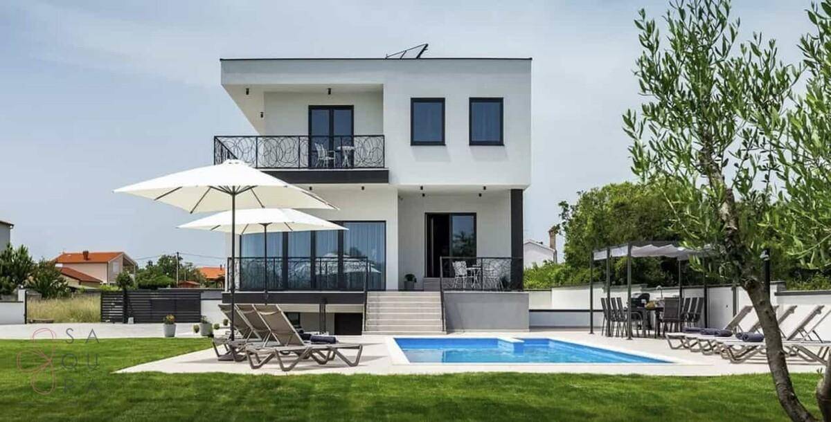 Frontansicht der modernen Villa mit Pool, Liegewiese und Sonnenschirmen an einem sonnigen Tag.