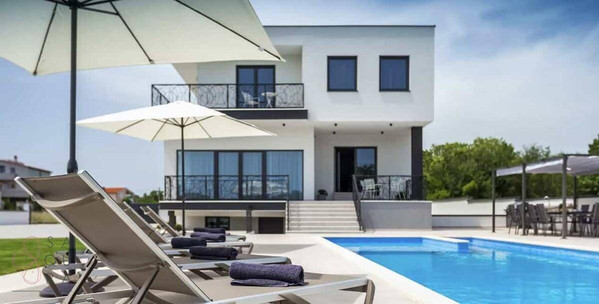 Blick auf die moderne Villa mit Poolbereich, Liegestühlen und Sonnenschirmen im Vordergrund.