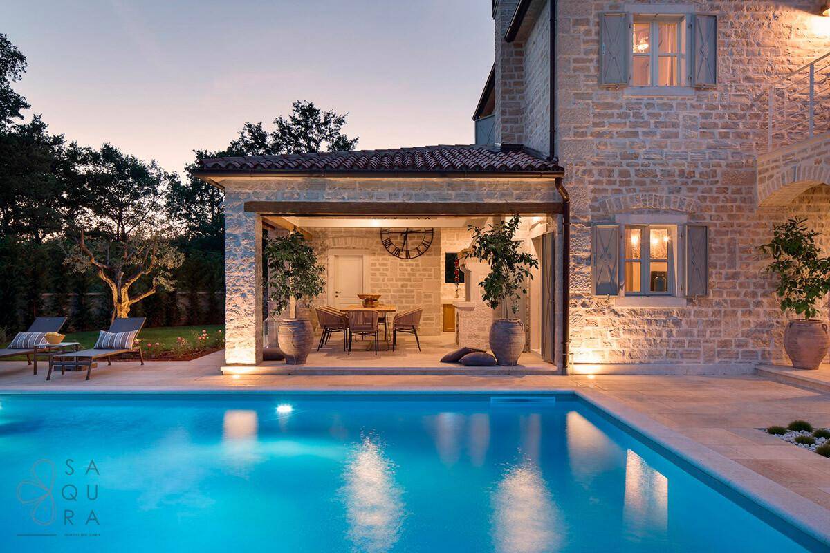 Beleuchteter Poolbereich und überdachte Terrasse mit Esstisch an der Steinfassade der Villa.