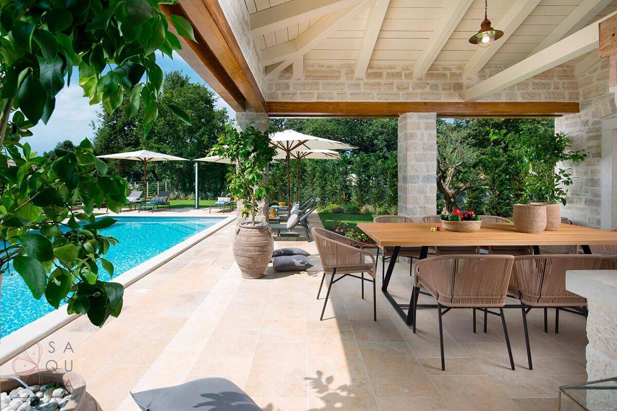 Überdachte Terrasse mit großem Esstisch und Blick auf den Pool und die grüne Gartenlandschaft.