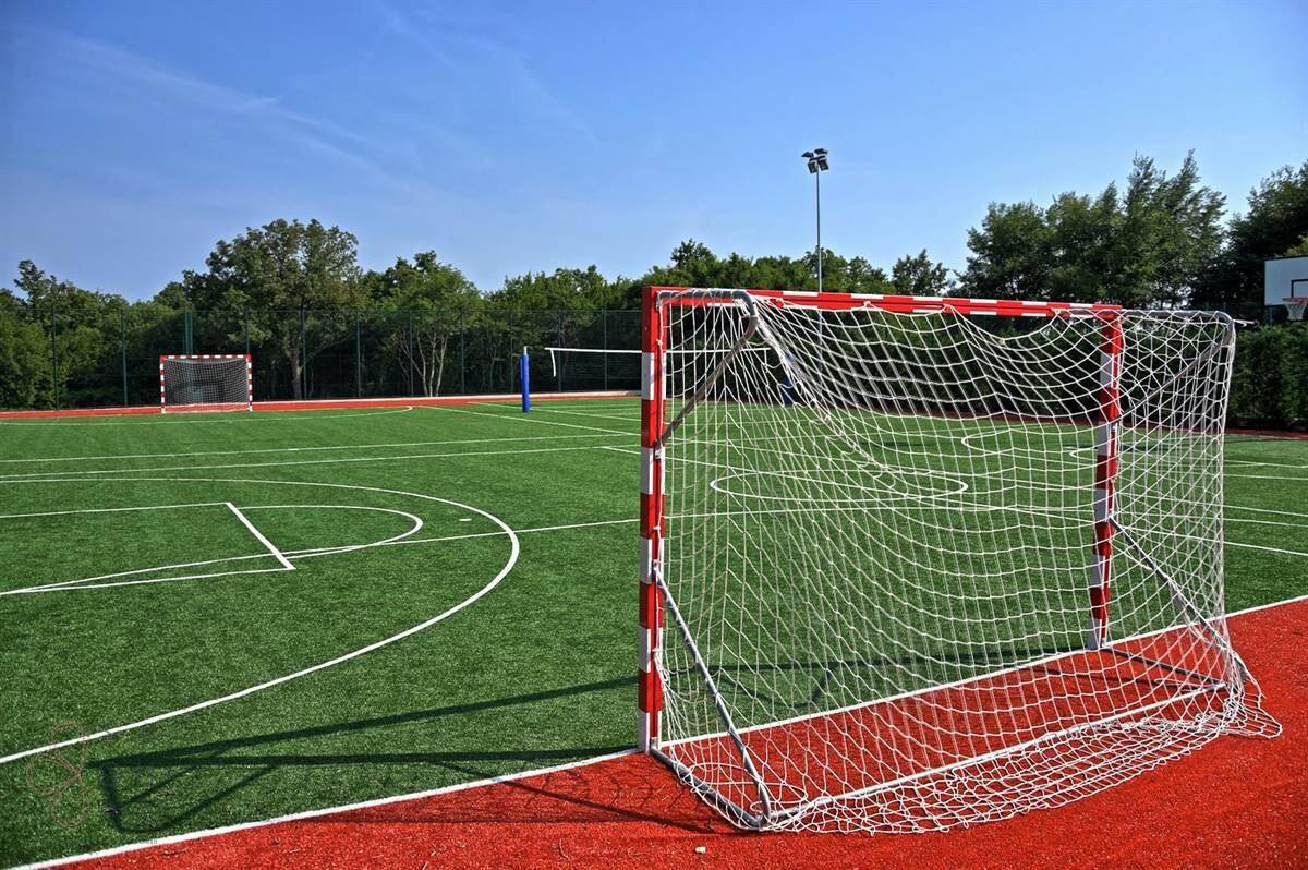 Multifunktionaler Sportplatz mit Fußballtoren und Volleyballnetz, umgeben von Bäumen.