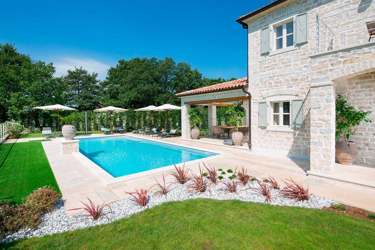 Gepflegter Gartenbereich mit Pool, mediterranen Pflanzen und Steinfassade der Villa unter blauem Himmel.