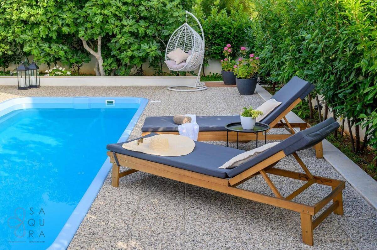 Sonniger Poolbereich mit Liegestühlen, Hängesessel und üppiger grüner Bepflanzung.