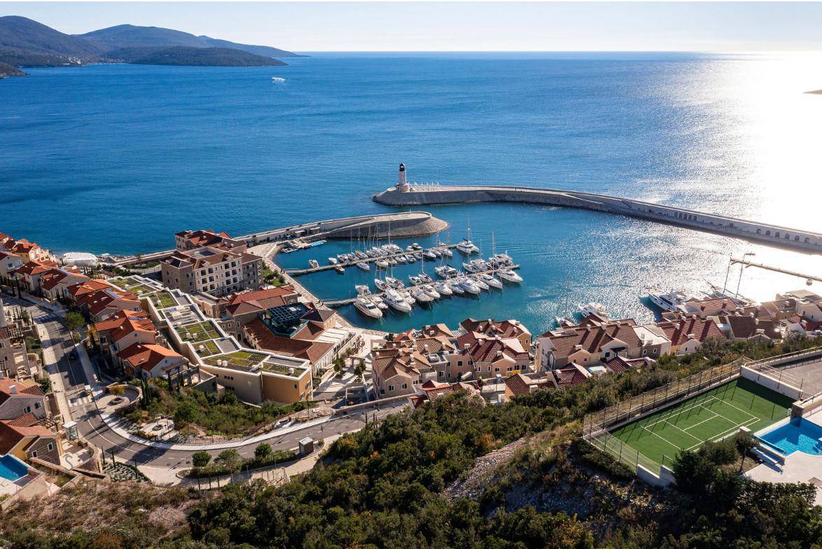 Luftaufnahme eines Yachthafens mit Leuchtturm, Küstenstadt und Tennisplatz am Meer.