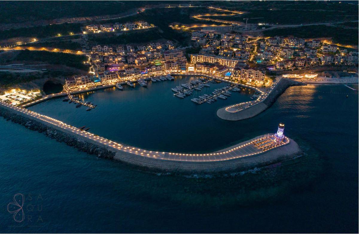 Nachtaufnahme des beleuchteten Hafens und der Küstensiedlung mit Leuchtturm und Yachten.