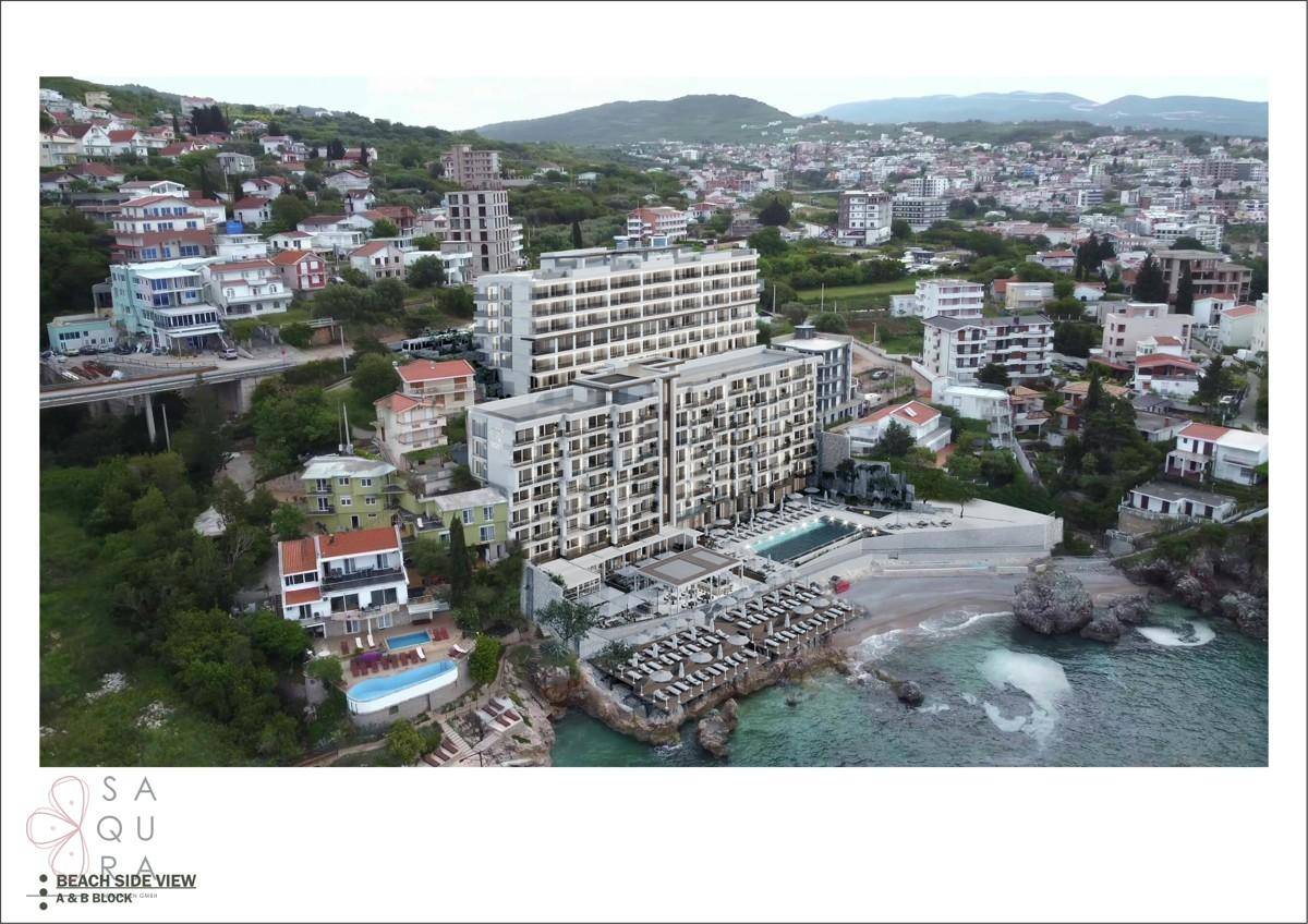 Luftaufnahme eines modernen Gebäudekomplexes direkt am Strand mit klarem Meerwasser und umliegender Bebauung.
