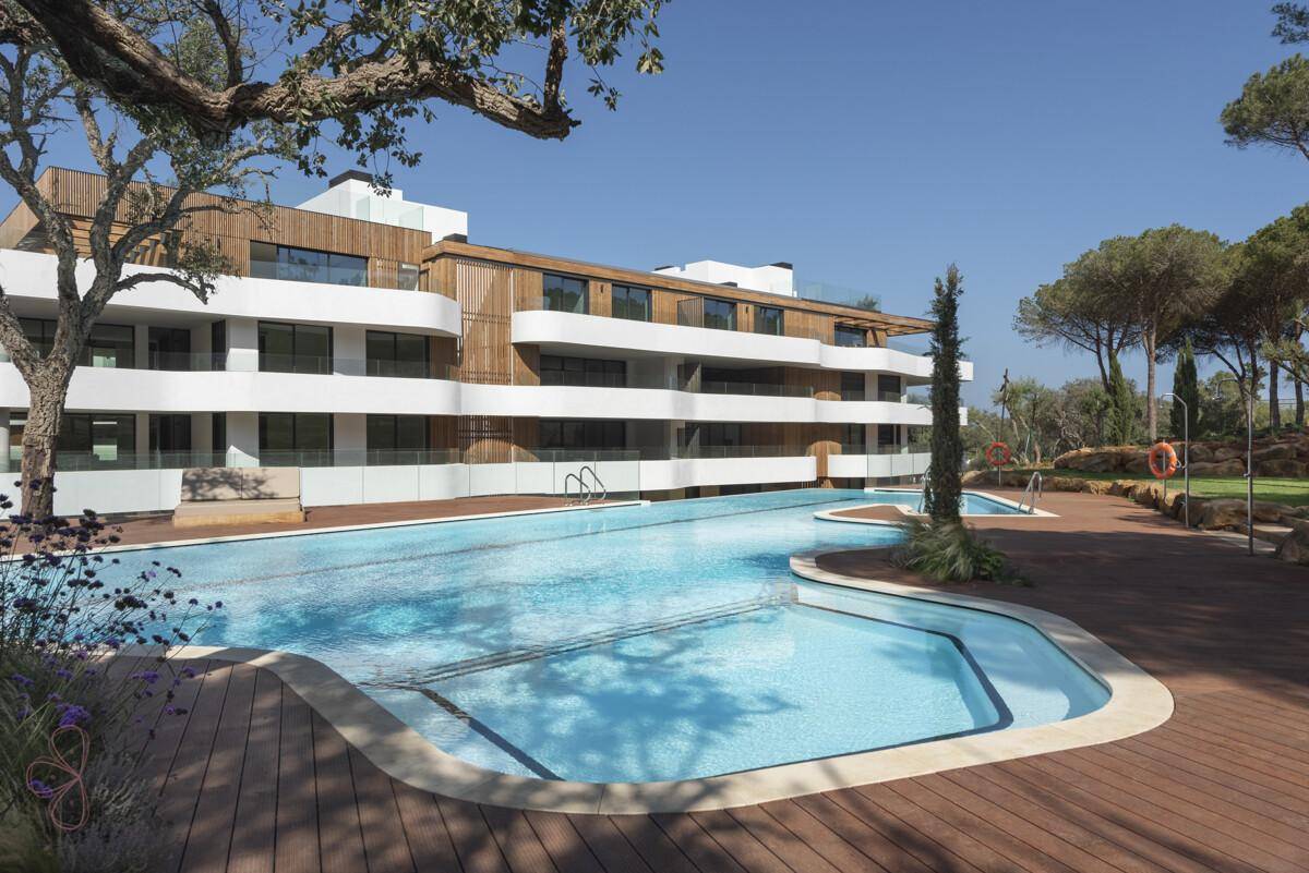 Weitläufiger Swimmingpool mit Holzdeck und Liegebereichen vor der modernen Wohnanlage unter blauem Himmel.