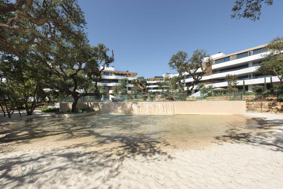 Künstlicher Strandbereich mit Wasserfall und üppiger Vegetation vor der modernen Wohnanlage.