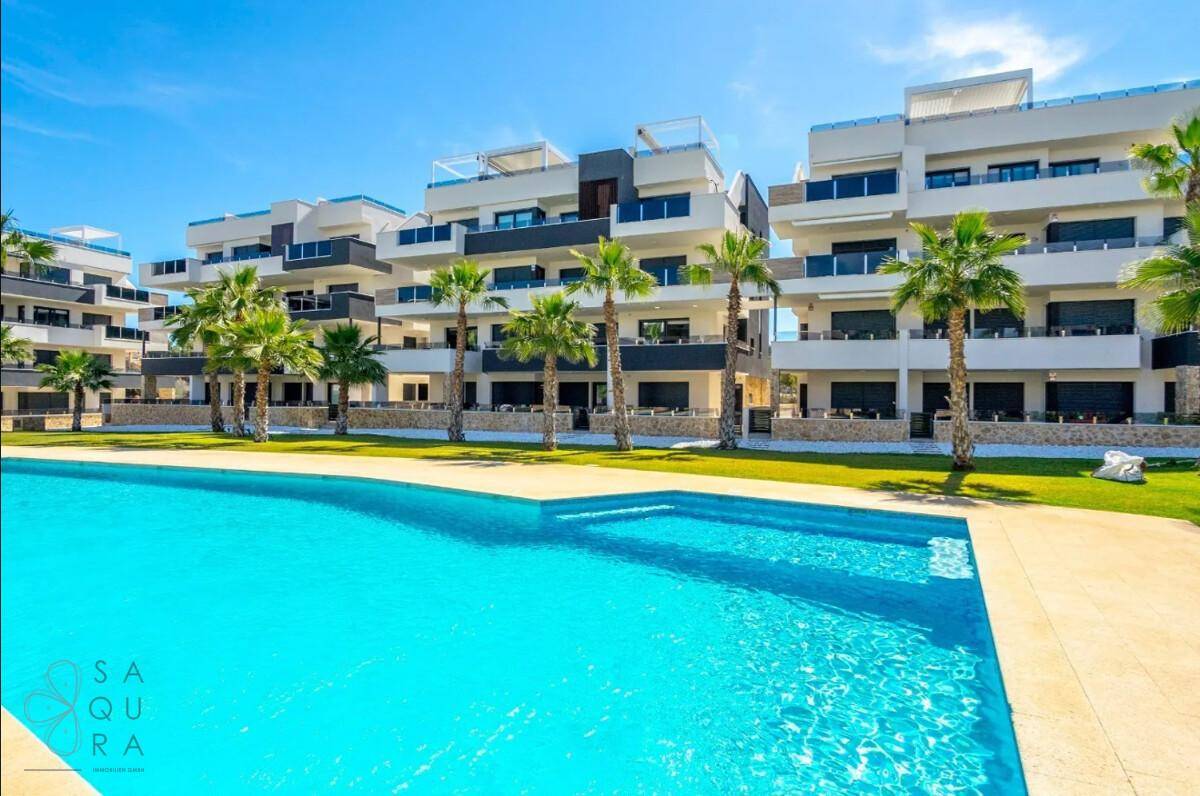 Weitläufige Poollandschaft vor den modernen Apartmentgebäuden unter strahlend blauem Himmel.