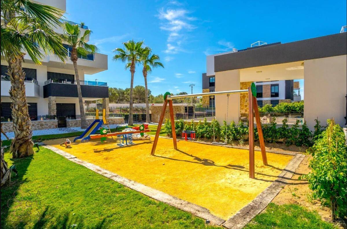 Moderner Kinderspielplatz mit Schaukeln und Rutsche, umgeben von Rasen und Palmen.