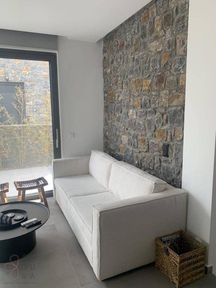 Wohnzimmer mit einer markanten Steinwand, modernem Sofa und großen Fenstern.