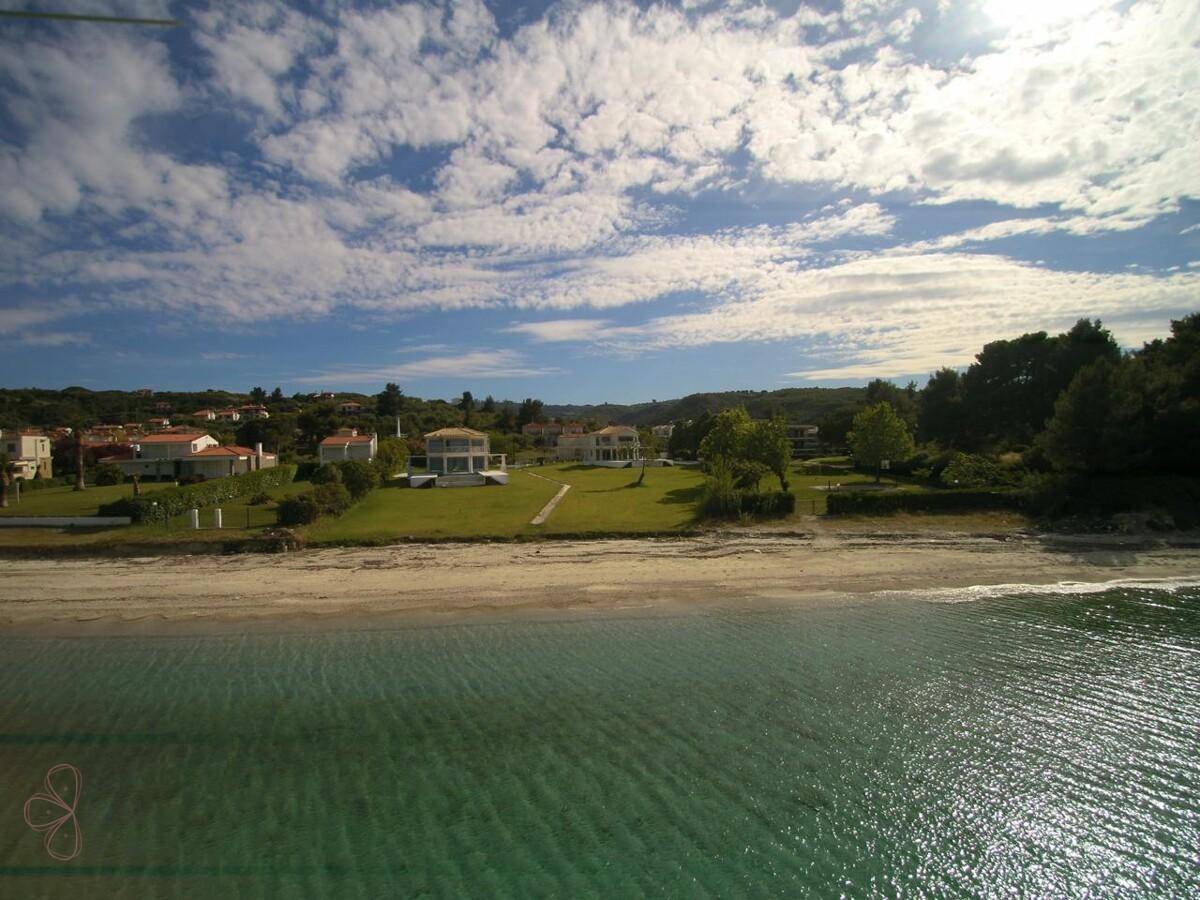 Weitläufiger Strandabschnitt mit klarem Wasser und Villen in der Ferne unter blauem Himmel.