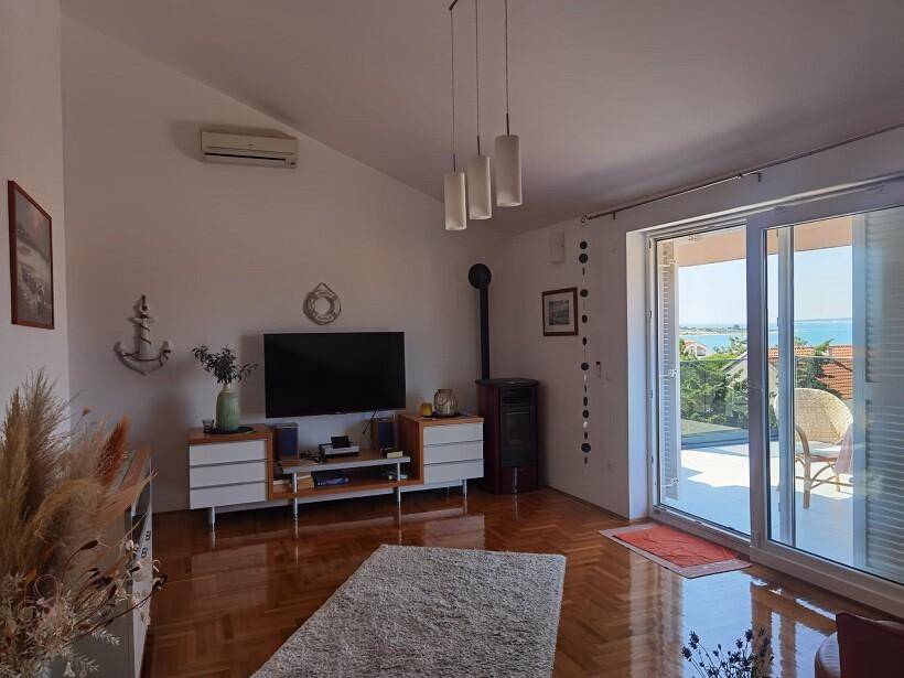 Wohnzimmer mit Parkettboden, Dachschräge, Holzofen und großen Fenstern zum Balkon mit Meerblick.