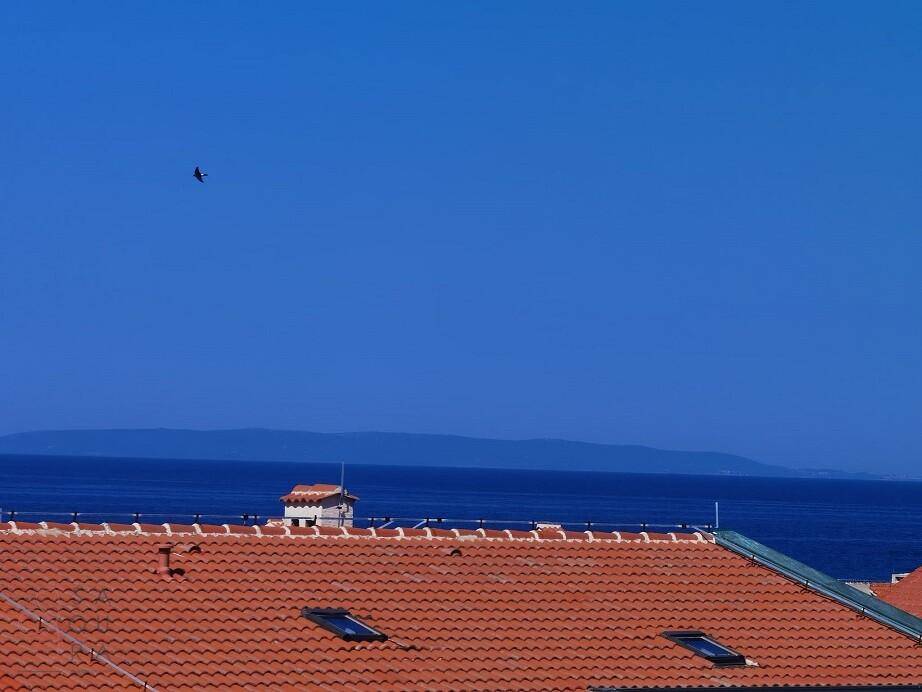 Blick über traditionelle rote Ziegeldächer auf das weite, blaue Meer und die ferne Küste.