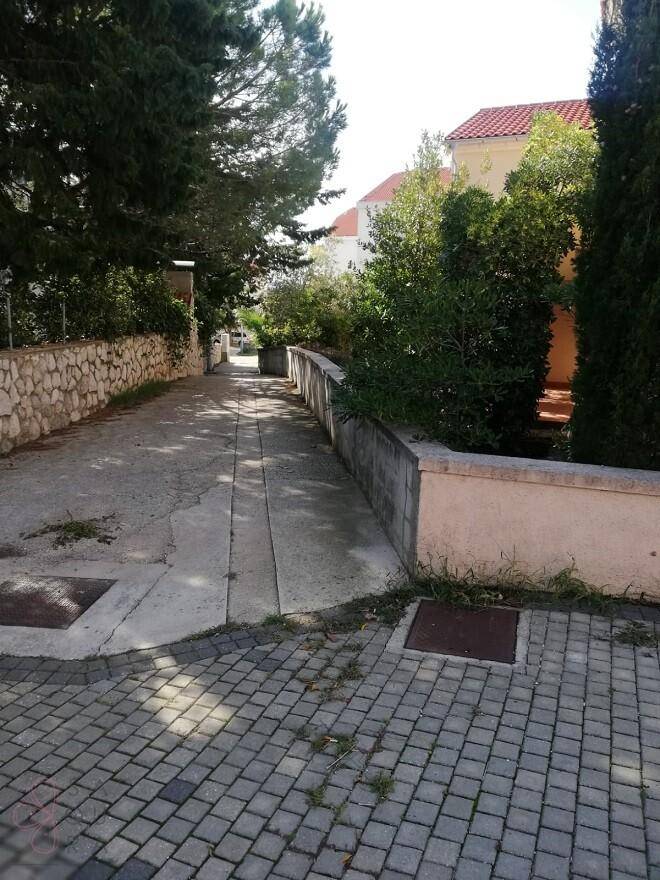 Gepflasterter Weg mit Steinmauer und üppiger Vegetation, der zu einem Gebäude führt.