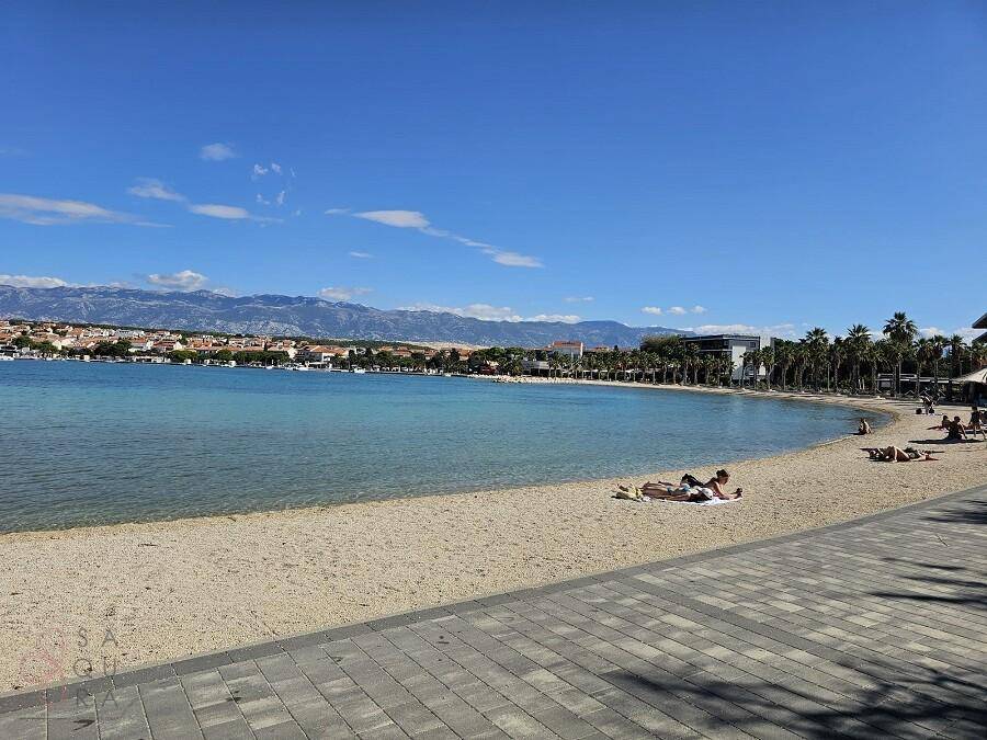 Weitläufiger Sandstrand mit azurblauem Meer und Bergen am Horizont unter strahlendem Himmel.