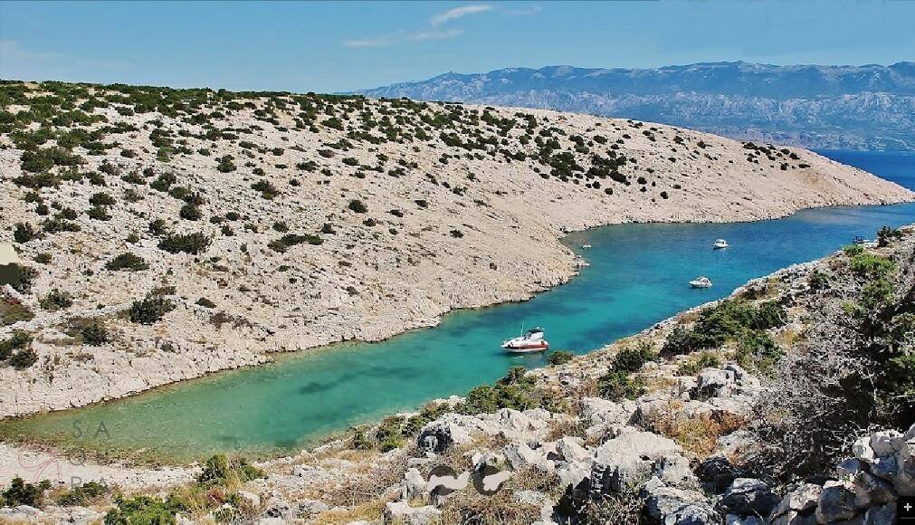 Idyllische Bucht mit türkisblauem Wasser und felsiger Küste unter strahlendem Himmel, einige Boote sichtbar.