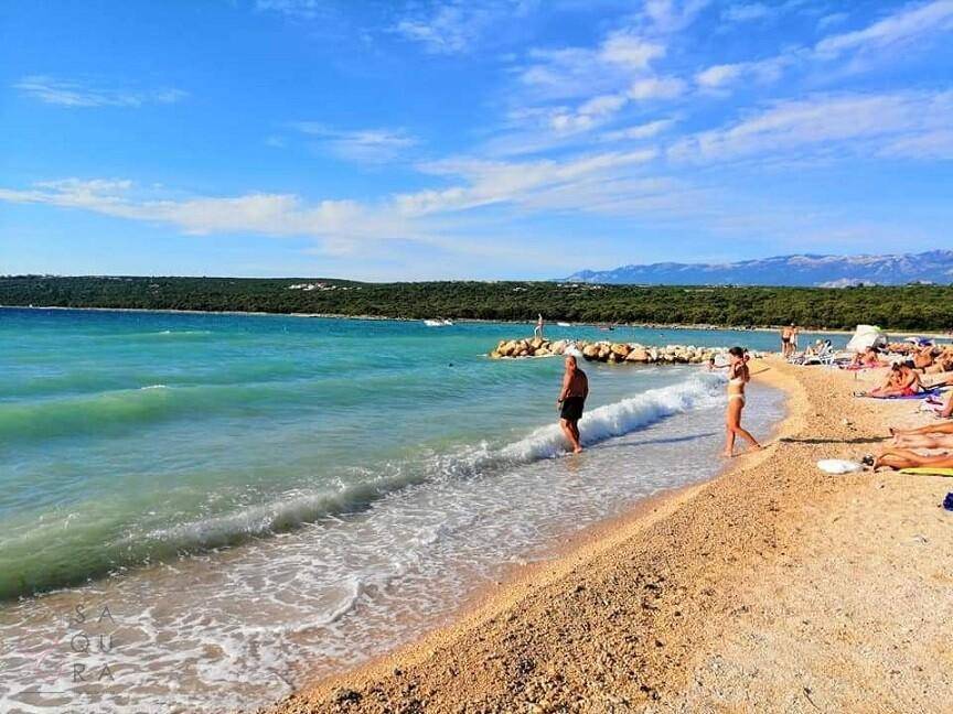 Belebter Strand mit Badegästen, sanften Wellen und Bergen im Hintergrund.