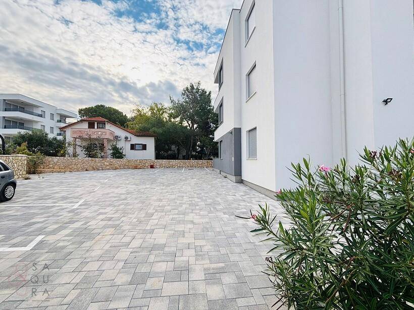 Außenansicht des modernen Gebäudes mit großem, gepflastertem Parkplatz und umliegender Bebauung.