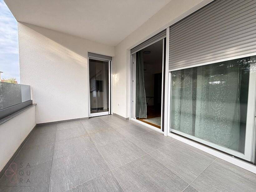 Heller Balkon mit grauen Fliesen, Glasgeländer und großen Schiebetüren zum Innenbereich.