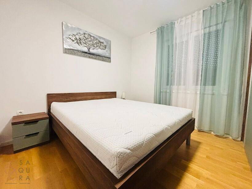 Schlafzimmer mit Holzbett, weißer Matratze, Nachttisch und hellgrünen Vorhängen am Fenster.