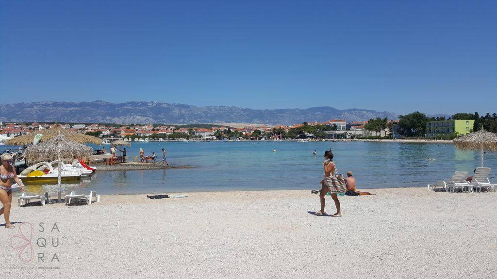 Sonniger Strand mit klarem blauen Wasser, Badegästen und Bergen am Horizont.