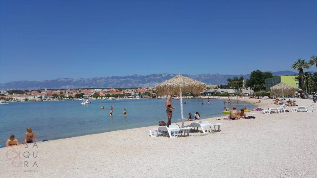 Weitläufiger Sandstrand mit Badegästen, Sonnenschirmen und dem Meer unter strahlend blauem Himmel.