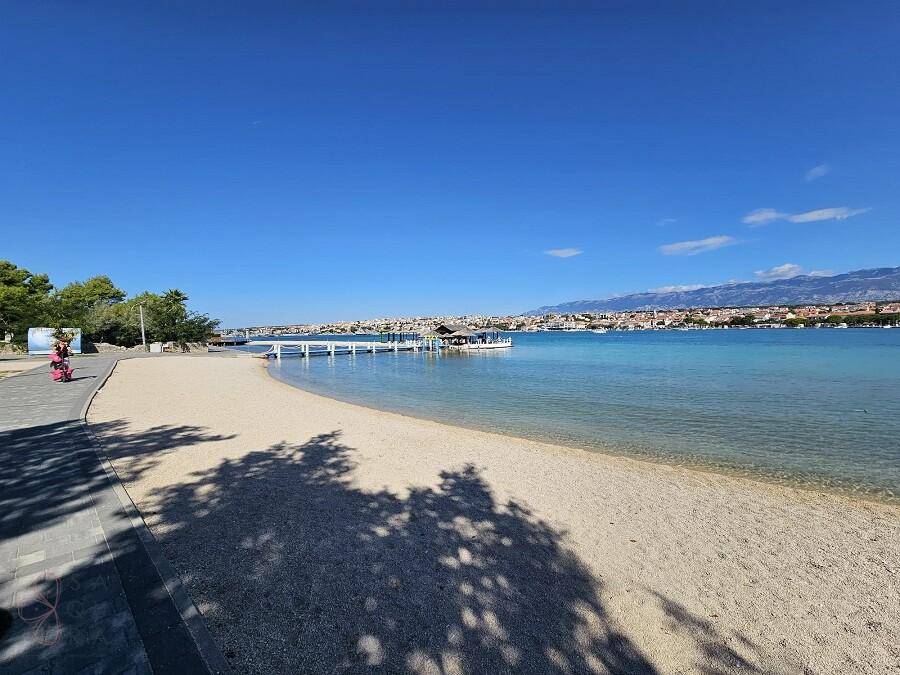 Sandstrand mit einem Steg, der ins klare blaue Wasser ragt, und einer Stadt in der Ferne.