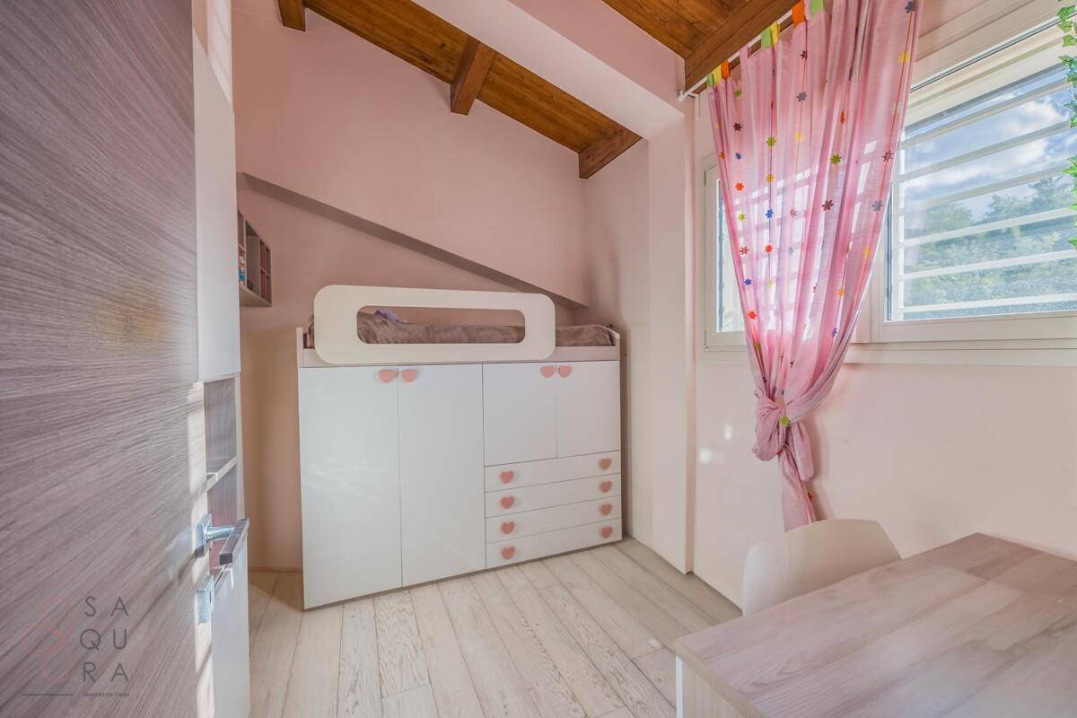 Freundliches Kinderzimmer mit rosa Vorhängen, Holzbalkendecke und einem Kinderbett über einem Schrank.