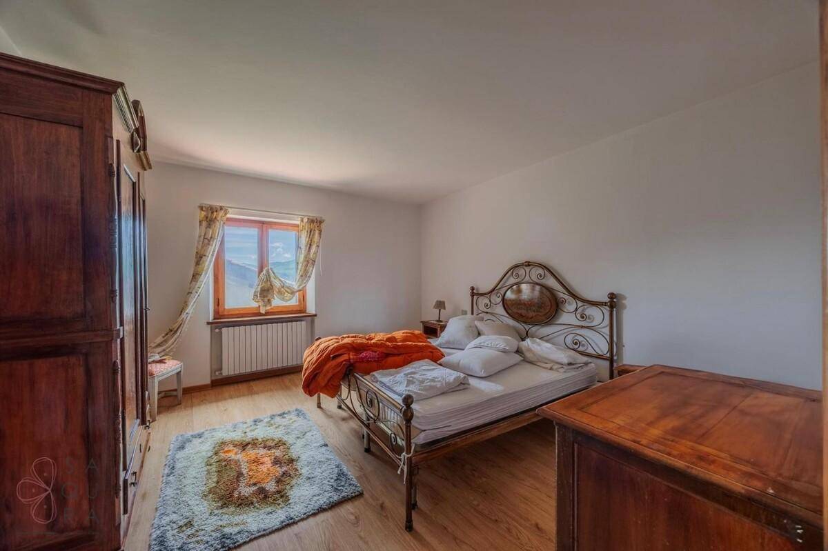Schlafzimmer mit schmiedeeisernem Bett, Holzschrank und hellem Holzboden, beleuchtet durch ein Fenster.