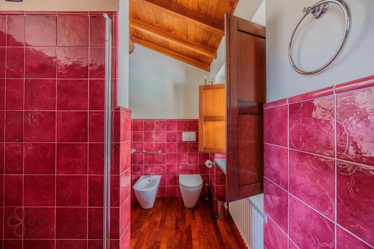 Badezimmer mit Duschkabine, Bidet, rotem Fliesensockel und Holzboden unter einer Holzbalkendecke.