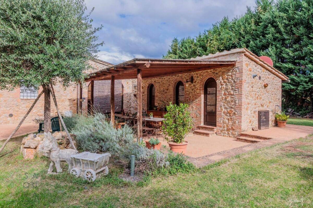 Überdachte Terrasse mit Holzbalken und Sitzgelegenheiten, umgeben von mediterranen Pflanzen und Rasen.