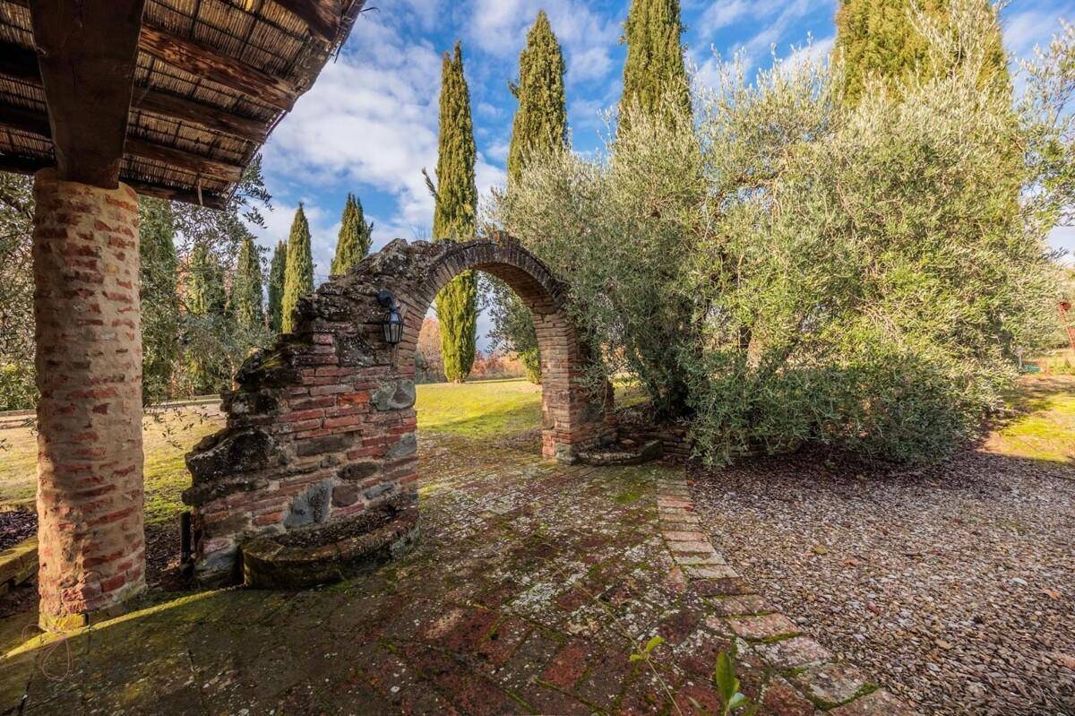 Historischer Steinbogen im Garten, umgeben von Olivenbäumen und Zypressen, mit Blick in die Ferne.