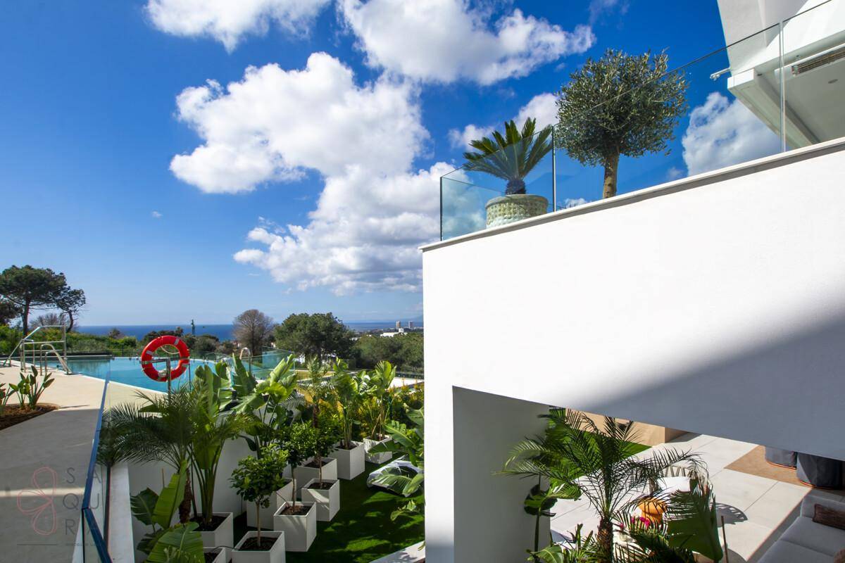 Blick von einem Balkon auf die Poolanlage, üppige Vegetation und das Meer in der Ferne.
