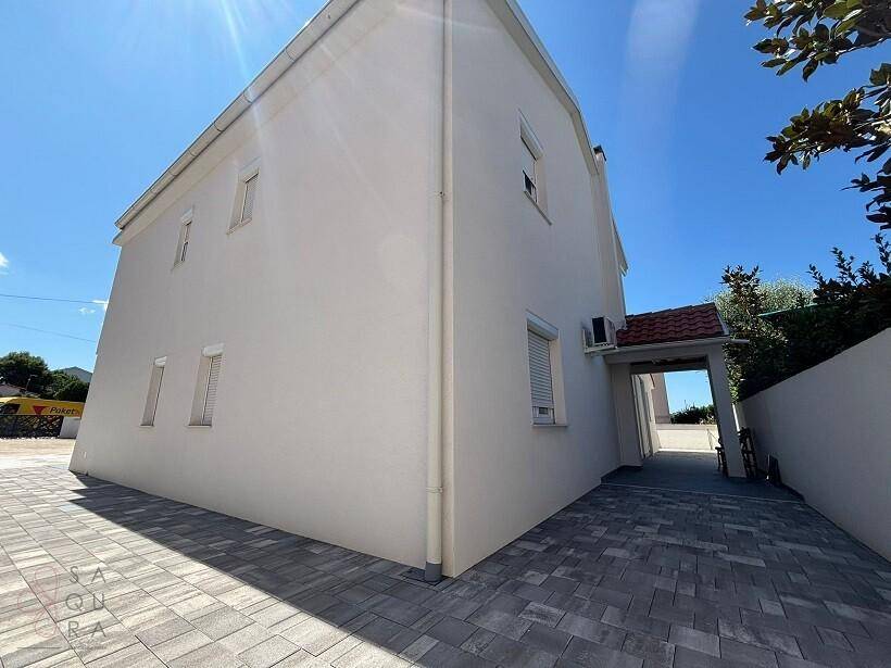 Seitenansicht des Hauses mit heller Fassade und gepflasterter Zufahrt, die zu einem überdachten Bereich führt.