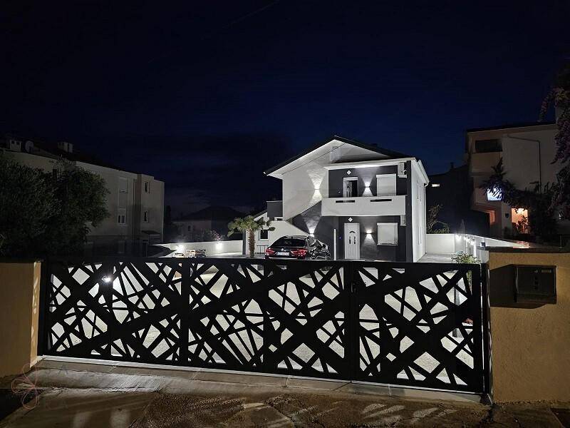 Modernes Haus bei Nacht, sichtbar hinter einem beleuchteten, dekorativen Metalltor mit geometrischem Muster.