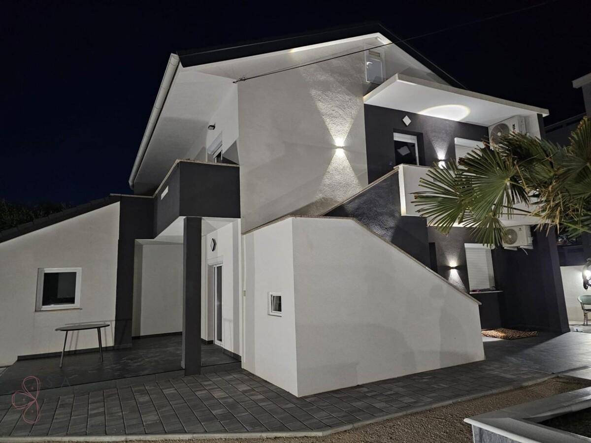 Modernes Haus bei Nacht mit beleuchteter Fassade, grauen Akzenten und gepflastertem Außenbereich.