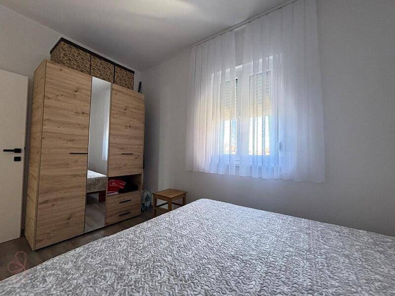 Schlafzimmer mit Holzoptik-Kleiderschrank, Doppelbett und einem Fenster mit hellem Vorhang.
