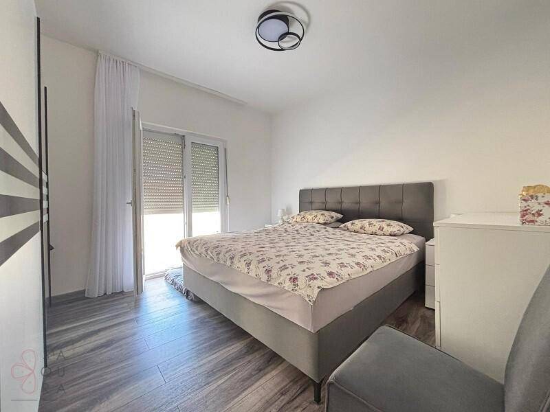 Modernes Schlafzimmer mit Doppelbett, Holzoptik-Boden und Zugang zu einem Balkon durch eine große Tür.
