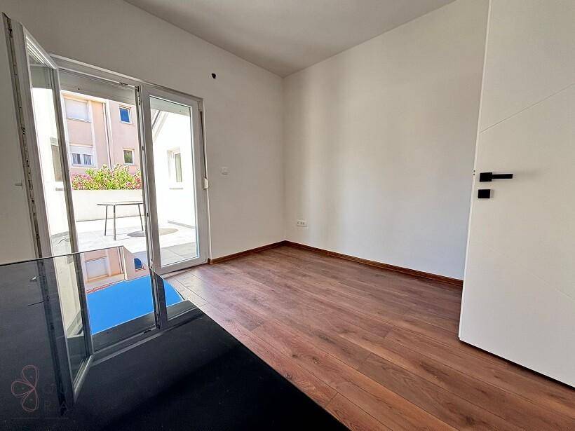 Leerer Raum mit Holzboden und großer Glasschiebetür, die Zugang zu einer sonnigen Terrasse bietet.