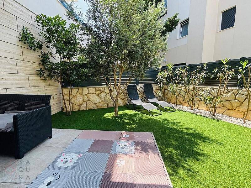 Sonniger Garten mit Kunstrasen, Olivenbaum und bequemen Liegestühlen für entspannte Stunden im Freien.