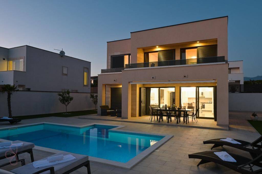 Moderne Villa bei Sonnenuntergang mit beleuchtetem Pool und gemütlichem Essbereich im Freien.
